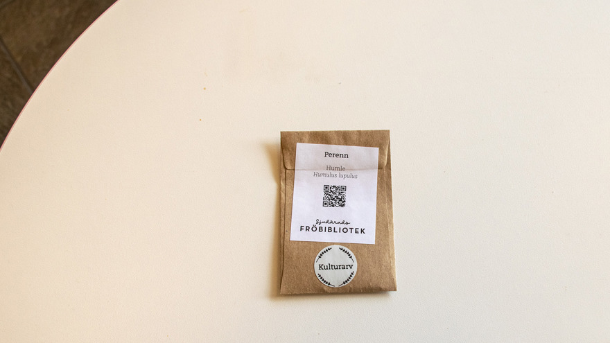 En brun liten fröpåse ligger på ett vitt bord. På den står det bland annat Humle, Perenn och Sjuhärads fröbibliotek. En QR-kod finns också på fröpåsen.