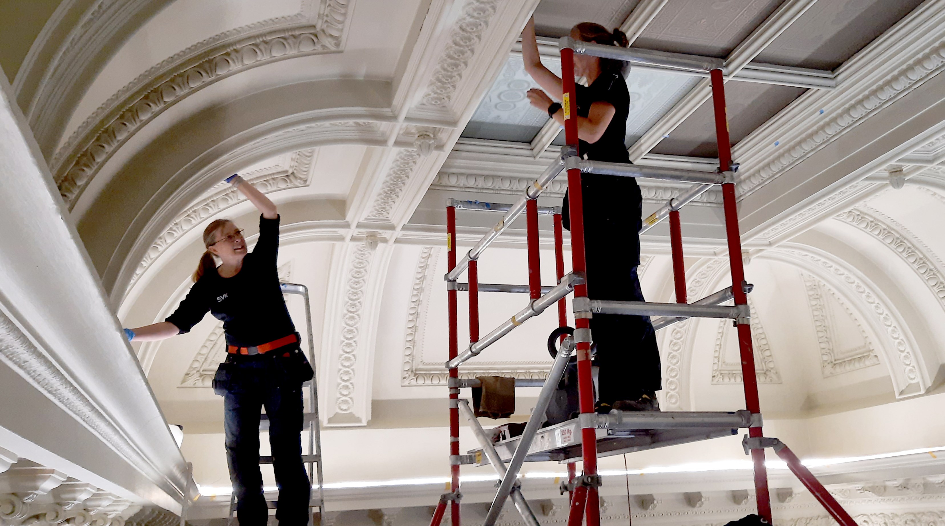 Två konservatorer står på en byggnadsställning och en stege och rengör ett tak.