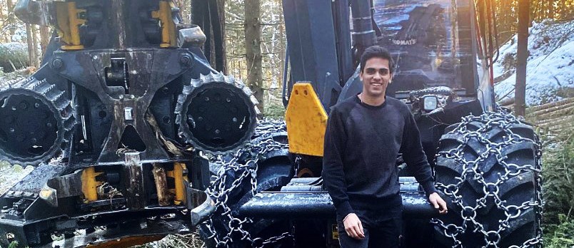 Oscar Elvingsson står i skogen med en skördare bakom sig