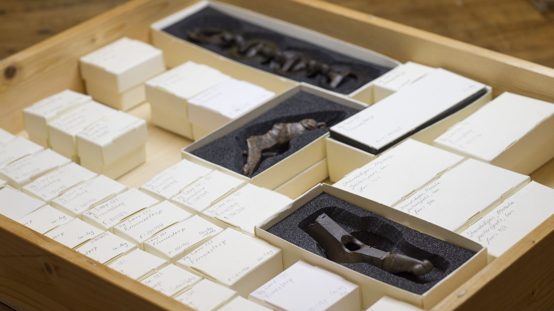 I en trälåda ligger mängder med små vita pappaskar. De flesta av askarna har lock, men några är öppna och där synd olika små arkeologiska föremål.