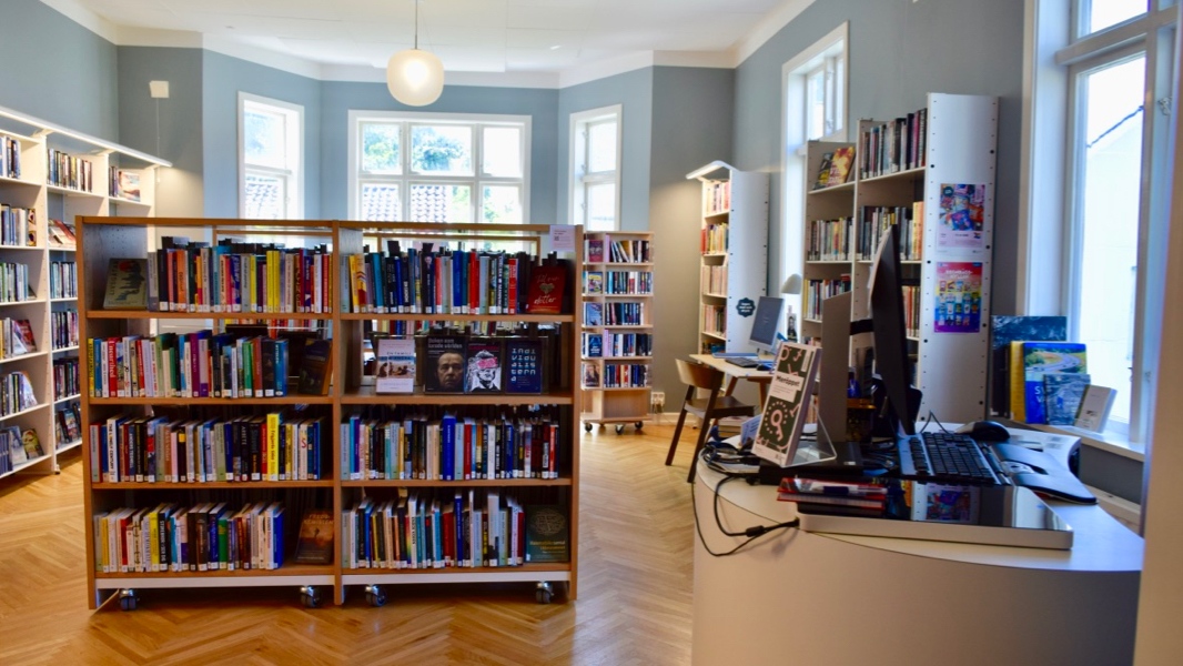 Södra Skärgårdens biblioteks vuxenavdelning. Rummet har ljusblå väggar och stora fönster i äldre stil. Rummet har bokhyllor både utmed väggarna och i mitten av rummet, till höger finns en utlåningsdisk.