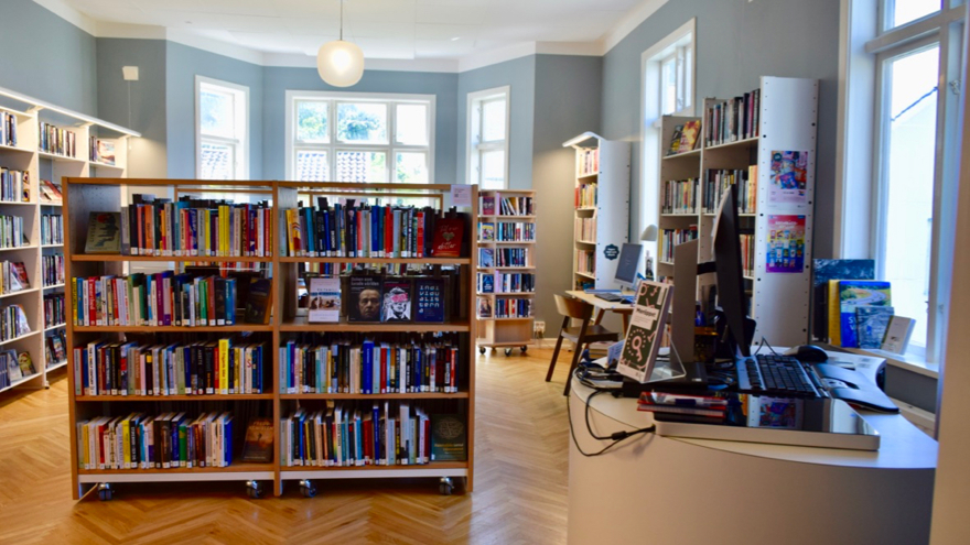 Södra Skärgårdens biblioteks vuxenavdelning. Rummet har ljusblå väggar och stora fönster i äldre stil. Rummet har bokhyllor både utmed väggarna och i mitten av rummet, till höger finns en utlåningsdisk.