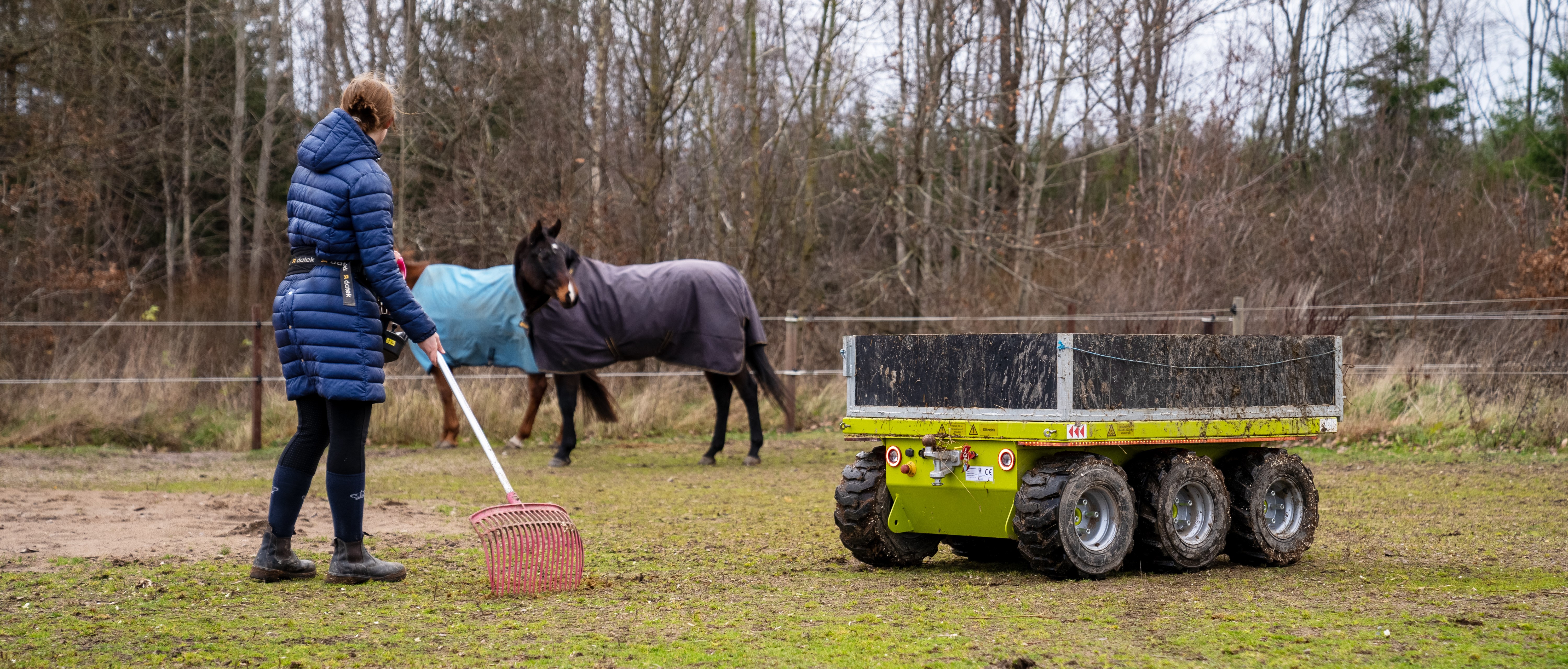 Stallhjälpredan står i en hästhage med två hästar i bakgrunden bredvid en kvinna med redskap i handen