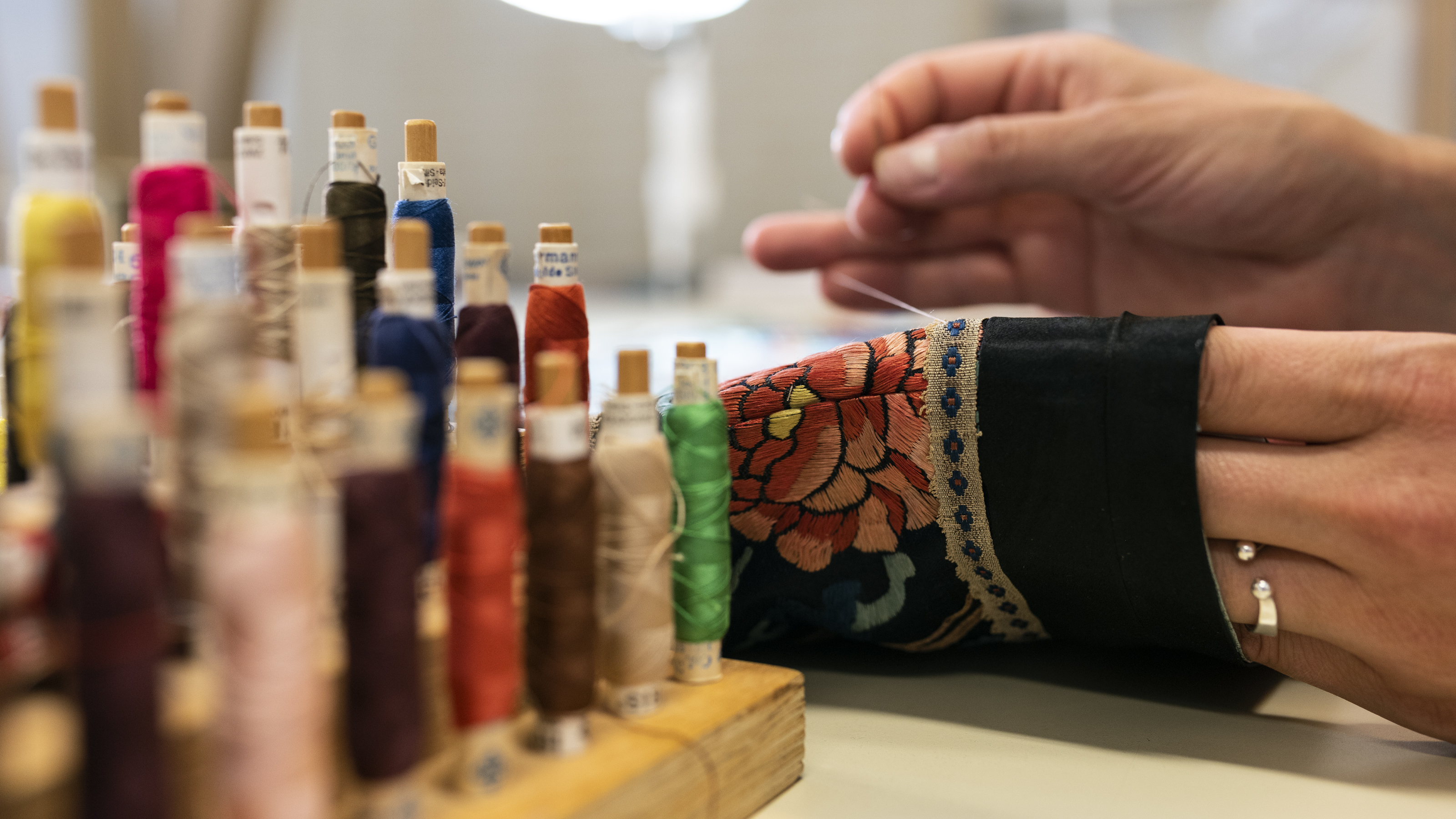 I förgrunden syns mängder med trådrullar i olika färger i ett ställ. Ett par händer syr på en svart ärm på ett plagg. Plagget har mängder med vackra broderier.